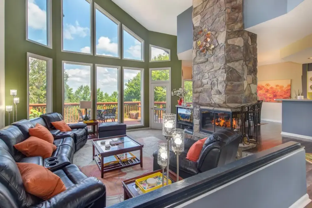 stone fireplace in the middle of a living room with black leather couches and open windows overlooking the mountains