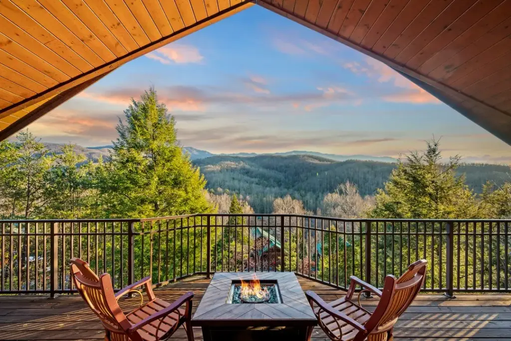 two wooden chairs with a firepit in the middle on a balcony overlooking the mountains and trees